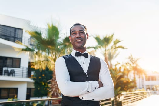 Posing for a camera. Black waiter in formal clothes is at his work outdoors at sunny daytime.