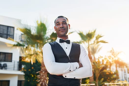 Posing for a camera. Black waiter in formal clothes is at his work outdoors at sunny daytime.