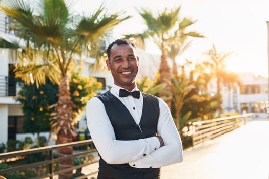 Posing for a camera. Black waiter in formal clothes is at his work outdoors at sunny daytime.