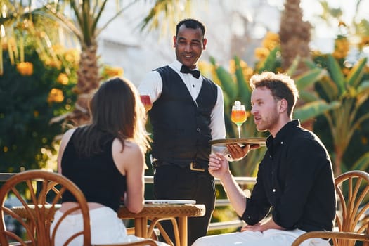 Serviced by waiter. Happy young couple is together on their vacation. Outdoors at sunny daytime.