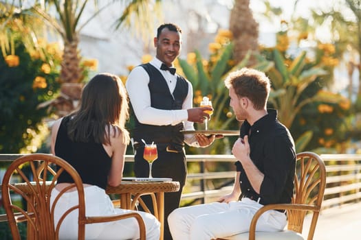 Serviced by waiter. Happy young couple is together on their vacation. Outdoors at sunny daytime.