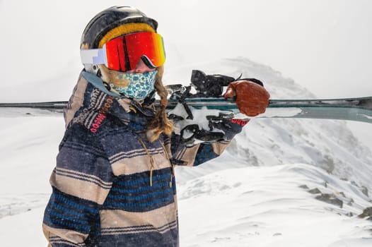 A girl in a jacket carries skis on her shoulder against the backdrop of mountains at a ski resort. Image of a woman with skis on her shoulder against the backdrop of a snowy hill.