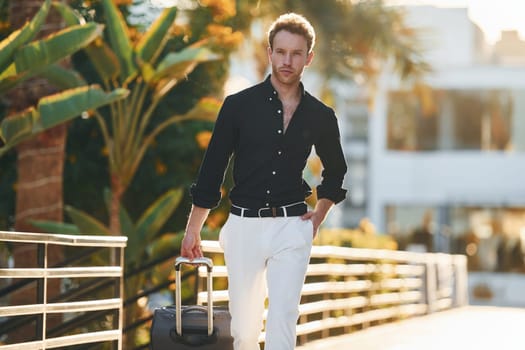 With travel bag. Young man is outdoors at sunny daytime. Concept of vacation.