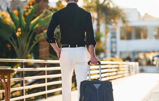 With travel bag. Young man is outdoors at sunny daytime. Concept of vacation.