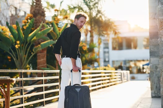 With travel bag. Young man is outdoors at sunny daytime. Concept of vacation.