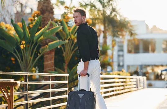 With travel bag. Young man is outdoors at sunny daytime. Concept of vacation.