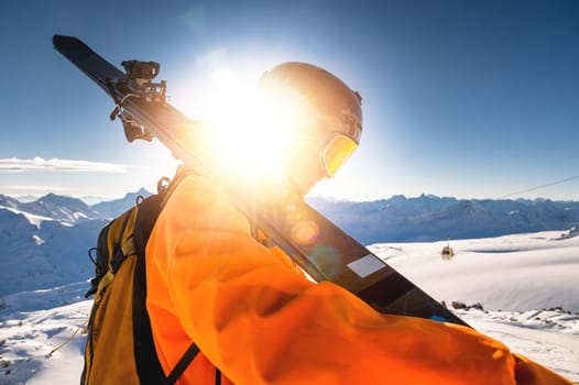The skier stands on the top of the mountain and enjoys the view of the beautiful winter mountains on a sunny day. Man holding skis on his shoulder.
