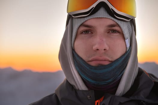 Portrait of a pensive skier in the mountains at sunset. A young man looks at the camera while standing in ski goggles against the backdrop of a sunset sky in the mountains.