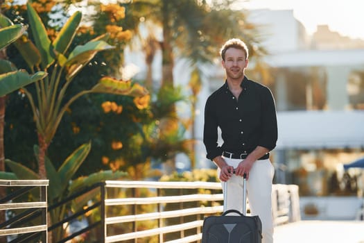 With travel bag. Young man is outdoors at sunny daytime. Concept of vacation.