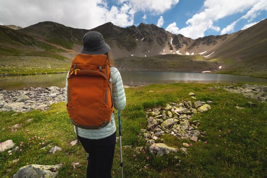 A traveling free man with an orange backpack walks along a path towards a high mountain range and a lake. Dramatic landscape with a tourist high in the mountains.