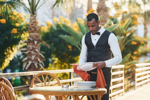 Wipes plates by red cloth. Black waiter in formal clothes is at his work outdoors at sunny daytime.