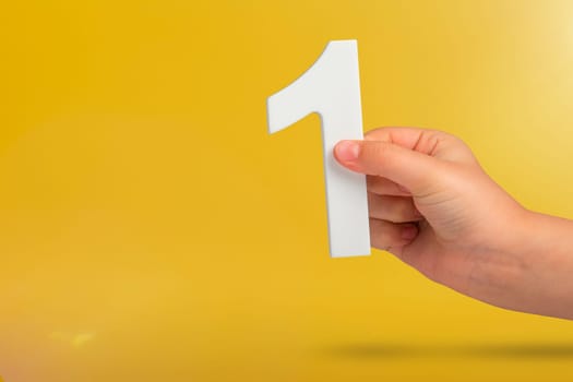 Number one in hand. A hand holds a white number one on a yellow background with copy space. Concept with number one. 1 percent rate, birthday, first or winner