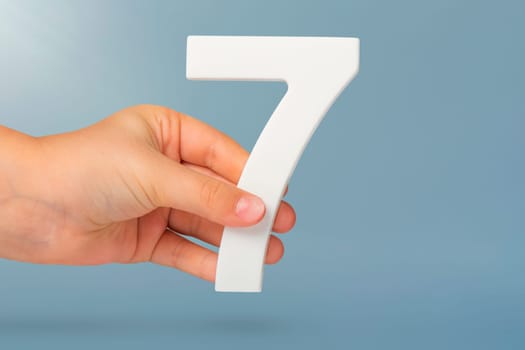 Number seven in hand. Hand holding white number 7 on blue background with copy space. Concept with number seven. Birthday 7 years, percent, seventh grade or day