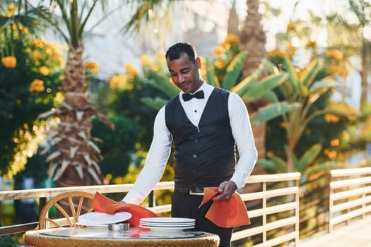 Wipes plates by red cloth. Black waiter in formal clothes is at his work outdoors at sunny daytime.