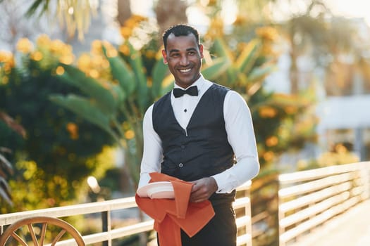 Wipes plates by red cloth. Black waiter in formal clothes is at his work outdoors at sunny daytime.