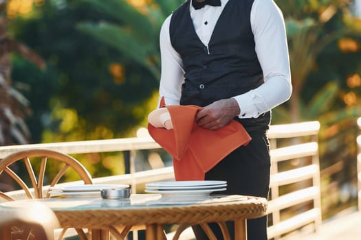 Wipes plates by red cloth. Black waiter in formal clothes is at his work outdoors at sunny daytime.