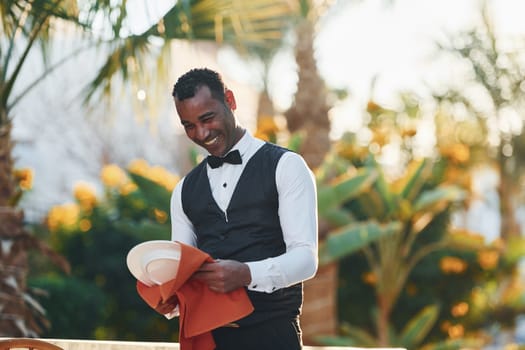 Wipes plates by red cloth. Black waiter in formal clothes is at his work outdoors at sunny daytime.