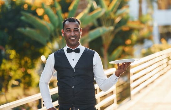 Holds empty plate. Black waiter in formal clothes is at his work outdoors at sunny daytime.