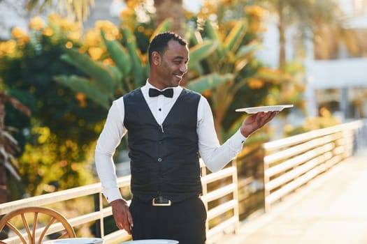 Holds empty plate. Black waiter in formal clothes is at his work outdoors at sunny daytime.