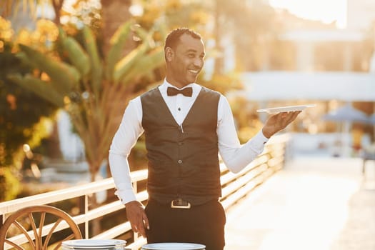 Holds empty plate. Black waiter in formal clothes is at his work outdoors at sunny daytime.