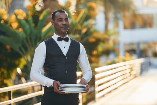 Holds empty plate. Black waiter in formal clothes is at his work outdoors at sunny daytime.
