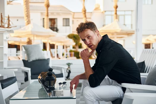 Sits by the table. Young man is outdoors at sunny daytime. Concept of vacation.