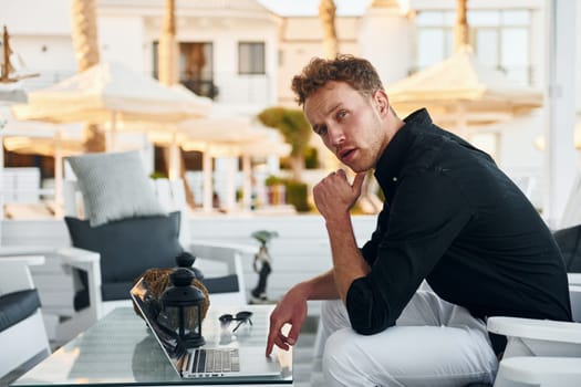 Sits by the table. Young man is outdoors at sunny daytime. Concept of vacation.