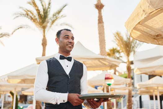 With order in hand. Black waiter in formal clothes is at his work outdoors at sunny daytime.