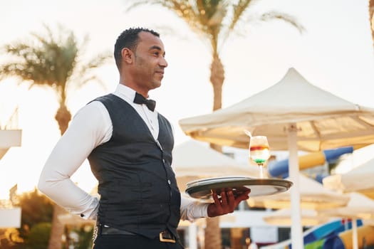 With order in hand. Black waiter in formal clothes is at his work outdoors at sunny daytime.