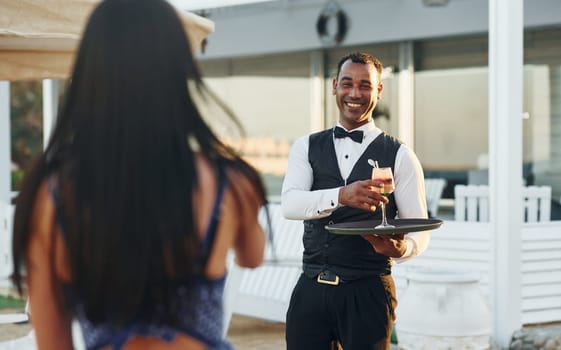 Order for woman. Black waiter in formal clothes is at his work outdoors at sunny daytime.