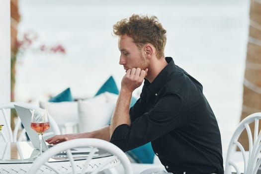 Sits by the table. Young man is outdoors at sunny daytime. Concept of vacation.