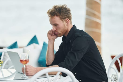 Sits by the table. Young man is outdoors at sunny daytime. Concept of vacation.