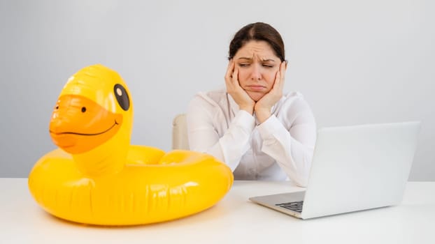 Caucasian woman sits at a table with a laptop and an inflatable duck on a white background. Office worker dreaming of vacation