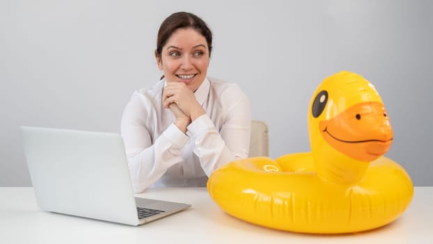 Caucasian woman sits at a table with a laptop and an inflatable duck on a white background. Office worker dreaming of vacation