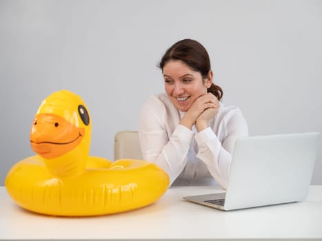 Caucasian woman sits at a table with a laptop and an inflatable duck on a white background. Office worker dreaming of vacation