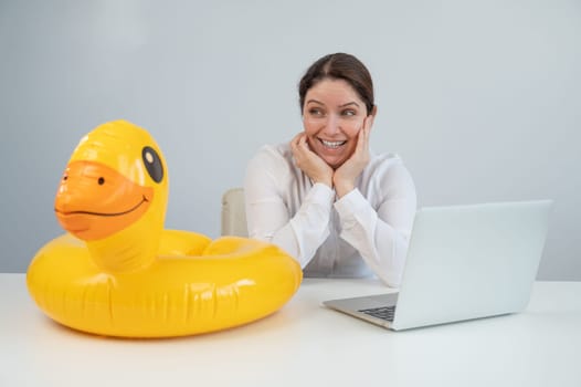 Caucasian woman sits at a table with a laptop and an inflatable duck on a white background. Office worker dreaming of vacation