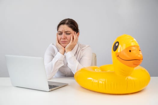 Caucasian woman sits at a table with a laptop and an inflatable duck on a white background. Office worker dreaming of vacation