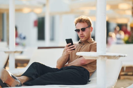 Sits and uses phone. Young man is outdoors at sunny daytime. Concept of vacation.