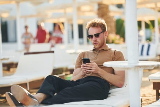 Sits and uses phone. Young man is outdoors at sunny daytime. Concept of vacation.
