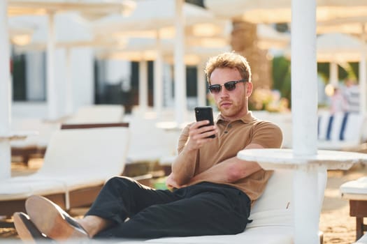 Sits and uses phone. Young man is outdoors at sunny daytime. Concept of vacation.