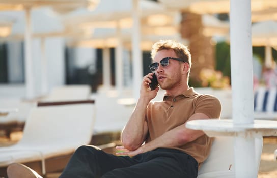Sits and uses phone. Young man is outdoors at sunny daytime. Concept of vacation.