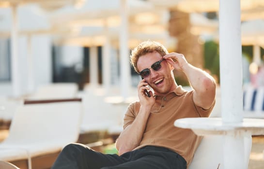 Sits and uses phone. Young man is outdoors at sunny daytime. Concept of vacation.