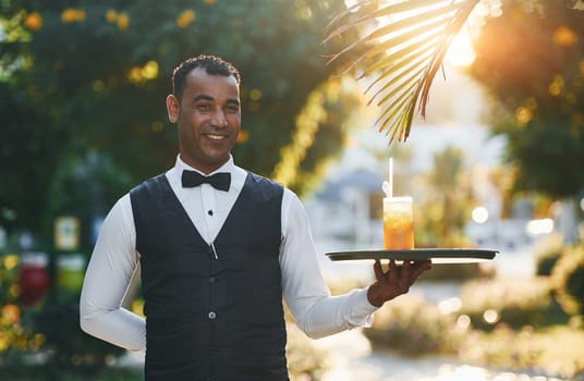 With order in hand. Black waiter in formal clothes is at his work outdoors at sunny daytime.