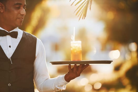 With order in hand. Black waiter in formal clothes is at his work outdoors at sunny daytime.