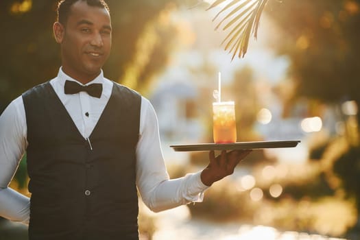 With order in hand. Black waiter in formal clothes is at his work outdoors at sunny daytime.