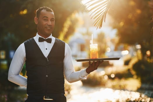 With order in hand. Black waiter in formal clothes is at his work outdoors at sunny daytime.