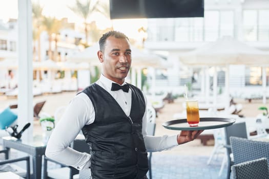 Black waiter in formal clothes is at his work outdoors at sunny daytime.