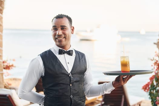 Black waiter in formal clothes is at his work outdoors at sunny daytime.