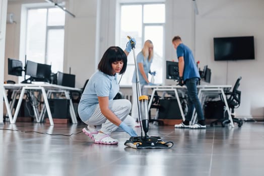Woman uses vacuum cleaner. Group of workers clean modern office together at daytime.