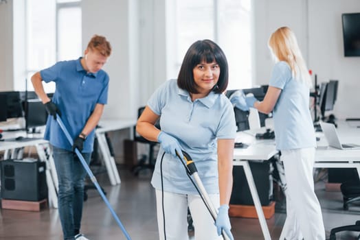 Woman uses vacuum cleaner. Group of workers clean modern office together at daytime.